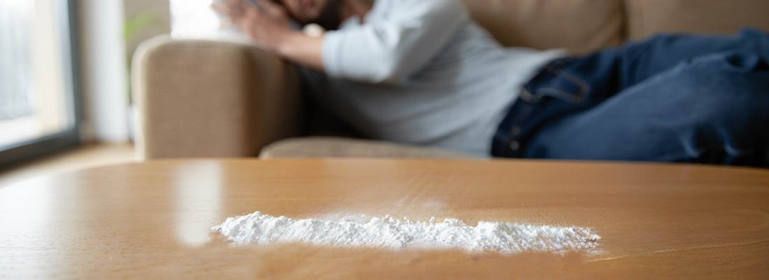 На столе белый порошок, сзади сиди мужчина на диване